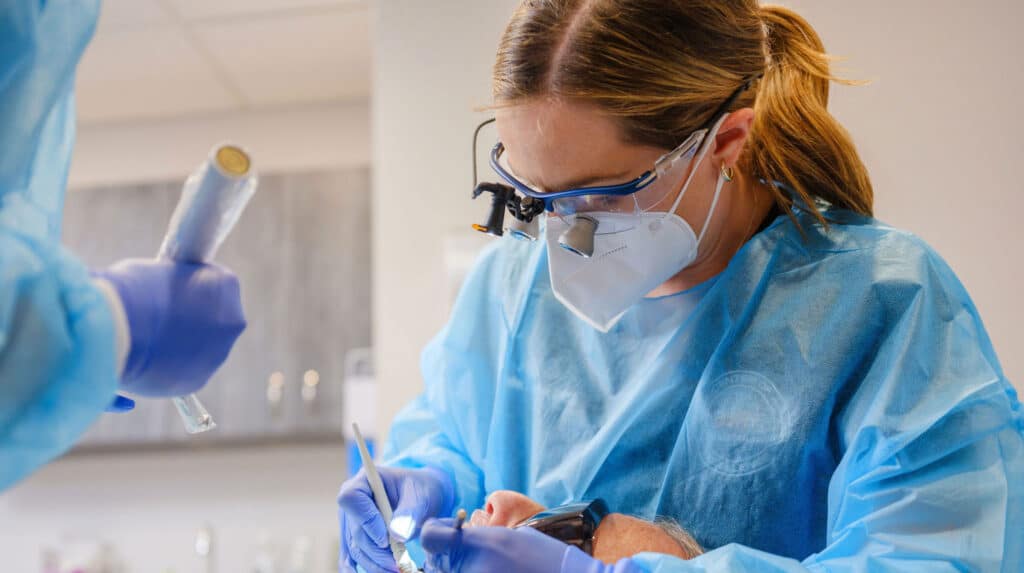 A female dentist wearing surgical loupes, a face mask, protective glasses, and blue gown performing a dental procedure on a patient, with an assistant holding equipment nearby.