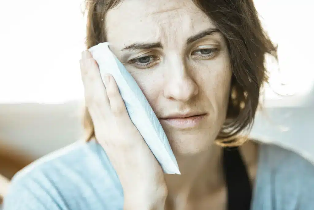 A woman holding an ice pack against her cheek, visibly expressing pain or discomfort due to a toothache, jaw pain, or post-dental surgery swelling.