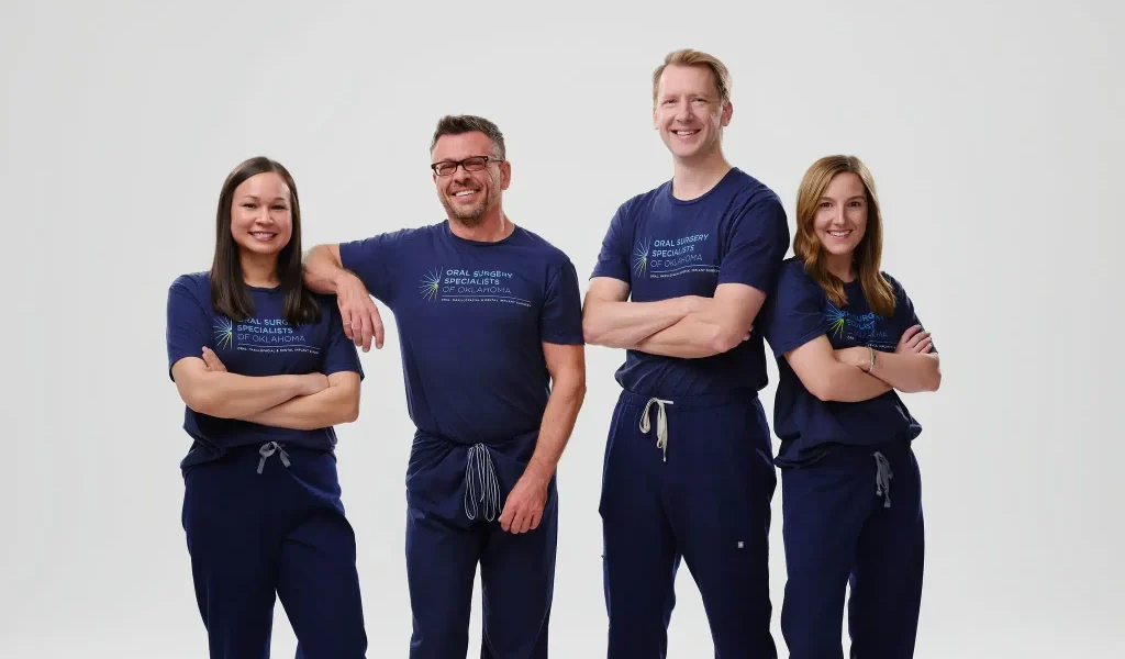 Four oral surgeons from Oral Surgery Specialists of Oklahoma posing together in matching navy branded T-shirts and scrub pants against a white studio background