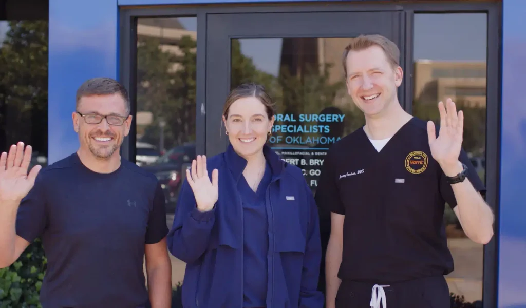 Three oral surgeons from Oral Surgery Specialists of Oklahoma smiling and waving outside the glass entrance of their medical office