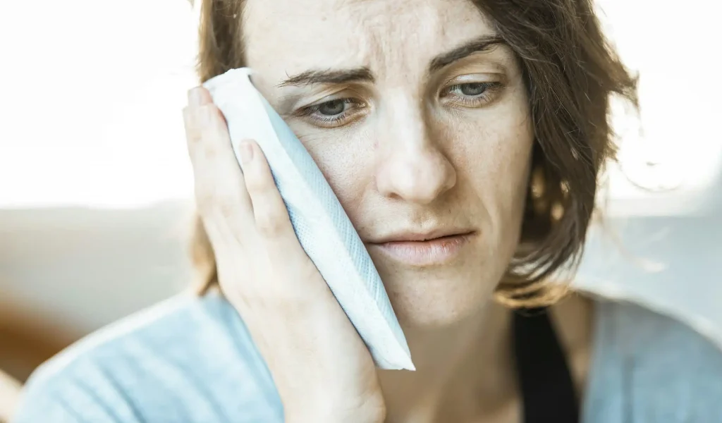 A woman holding an ice pack against her cheek, visibly expressing pain or discomfort due to a toothache, jaw pain, or post-dental surgery swelling.